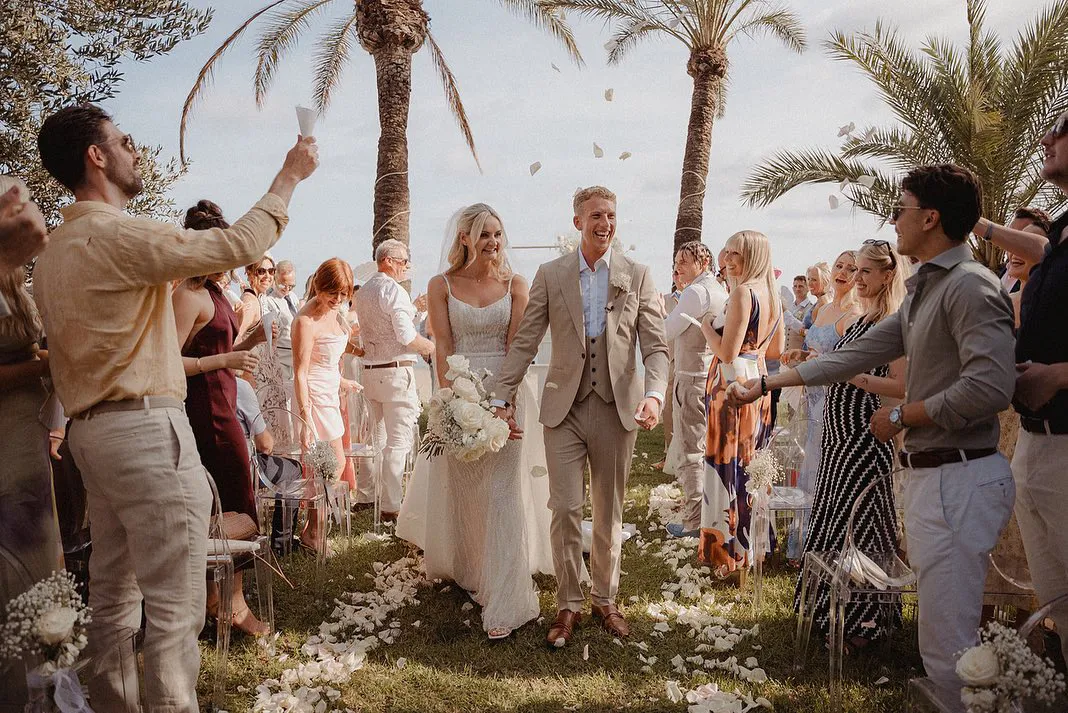 Bride and groom walking hand in hand down an outdoor aisle while guests throw flower petals, surrounded by palm trees.