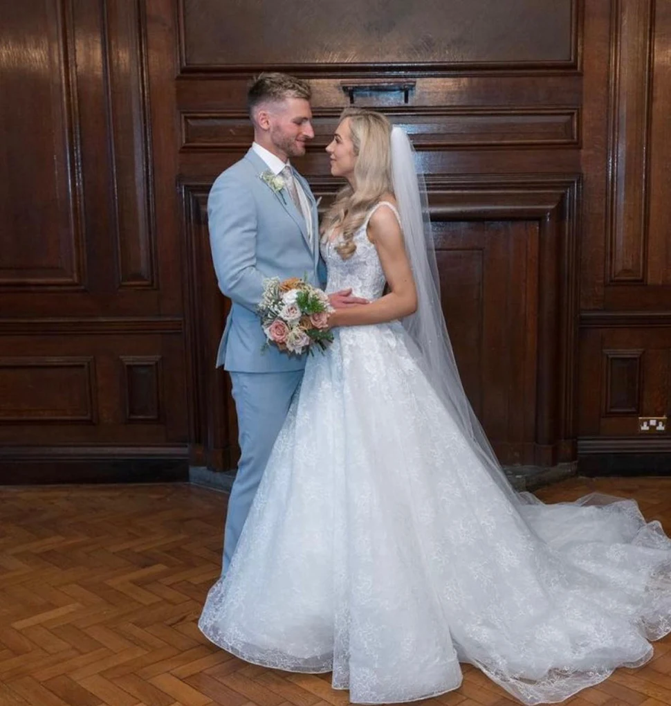 Bride in a white lace wedding gown and veil holding a bouquet, facing her groom in a light blue suit, standing on wooden parquet floor with dark wood panel walls.
