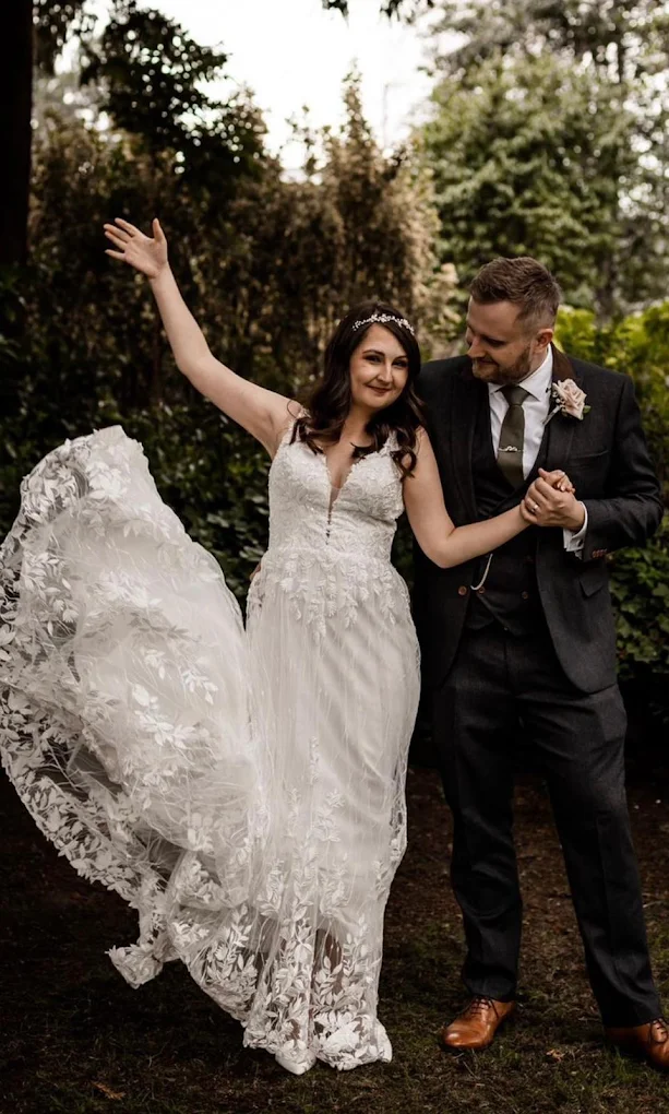 Bride in a flowing white lace wedding dress joyfully holding hands with the groom in a dark suit and brown shoes in an outdoor garden setting.