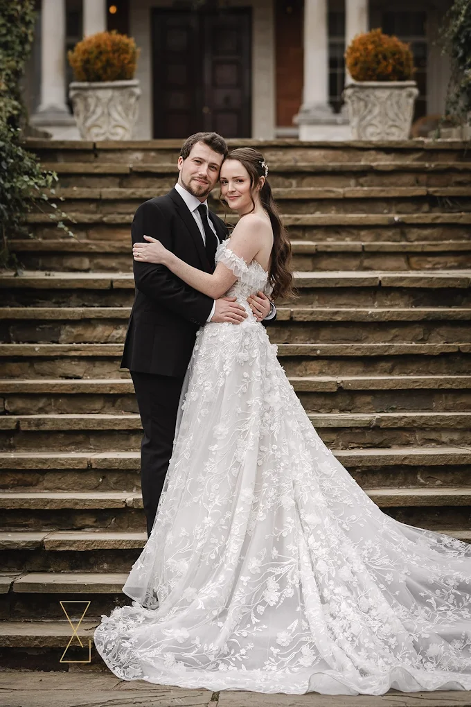 Bride and groom embracing on outdoor stone steps, bride wearing a detailed white lace gown with a long train.