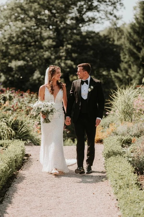 Bride in white dress holding bouquet and groom in tuxedo walking hand in hand on a garden path.