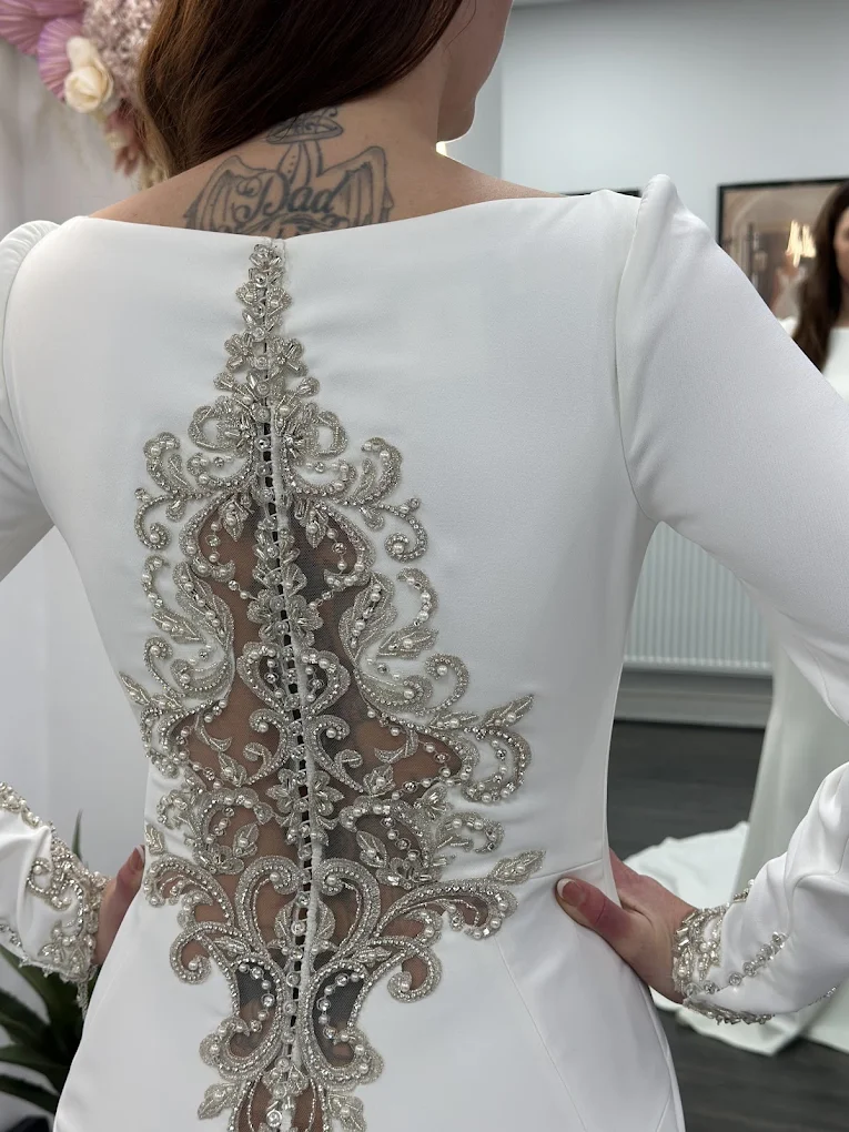 Back view of a woman wearing a white dress with intricate silver bead embroidery and sheer fabric details, showing a tattoo on her upper back.
