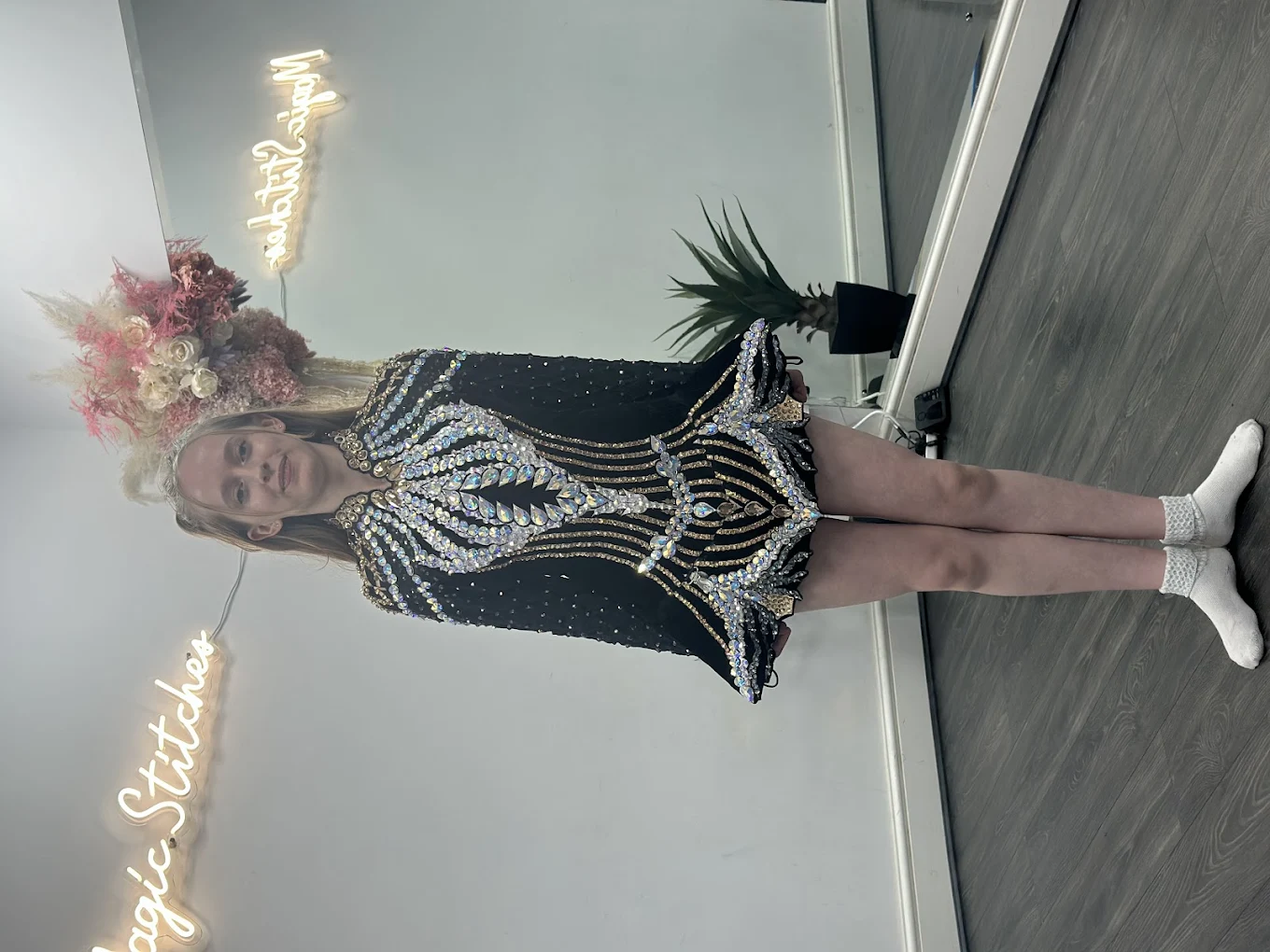 Woman wearing a black, gold, and silver sequined dress standing in a room with hardwood floors and a neon sign reading 'Magic Stitches' on the wall.