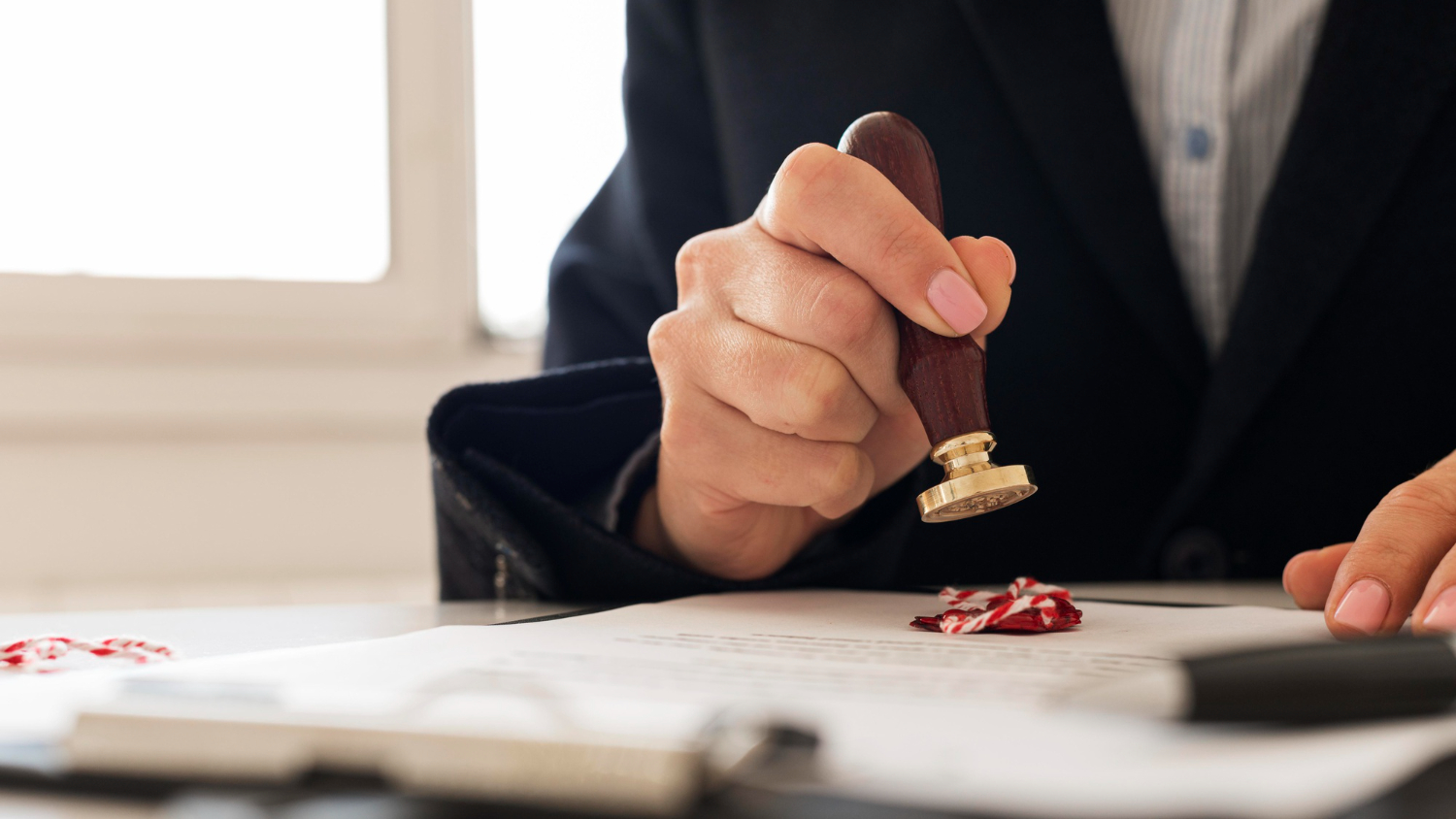 Person in dark suit pressing a wax seal stamp onto a document on a table.