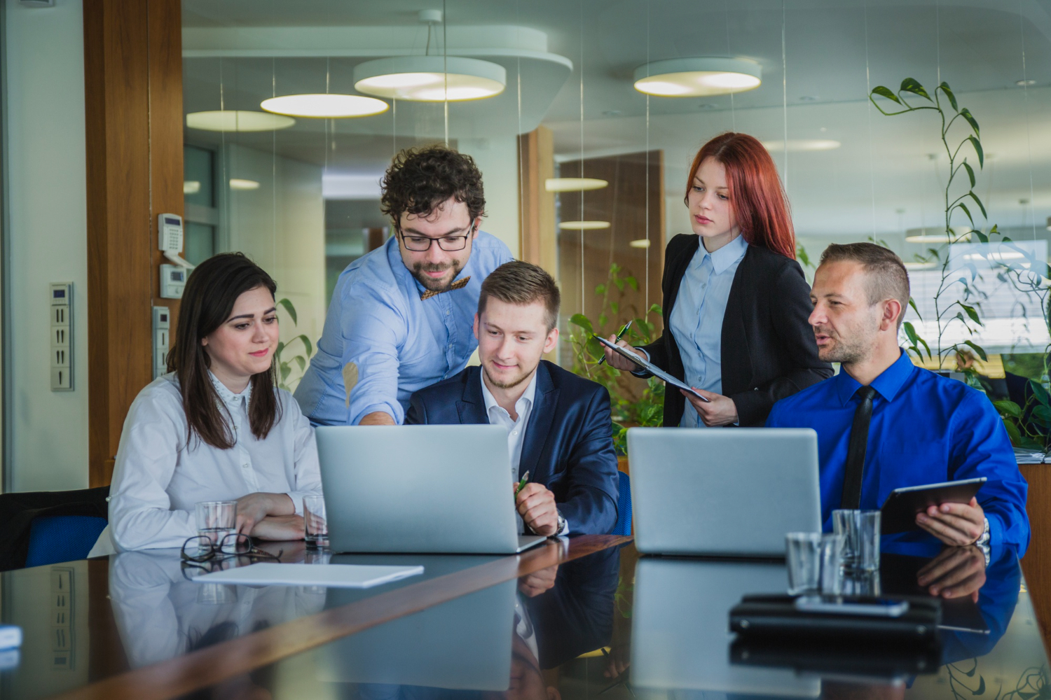 Five diverse professionals in business attire collaborating around a conference table with laptops in a modern office.