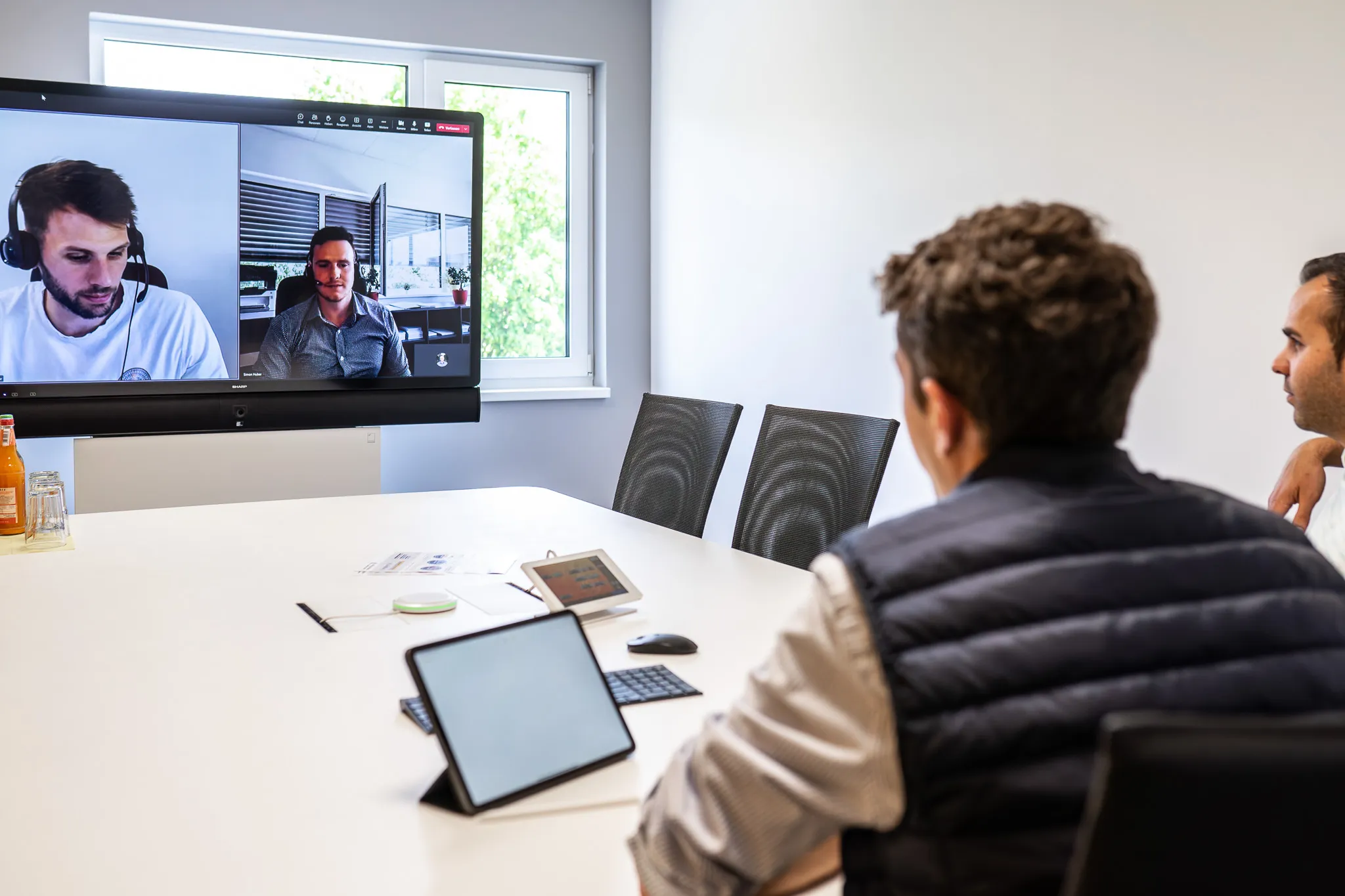Zwei Männer, die in einem kleinen Besprechungsraum ein hybrides Meeting abhalten, das auf einem großen Display anzeigen.