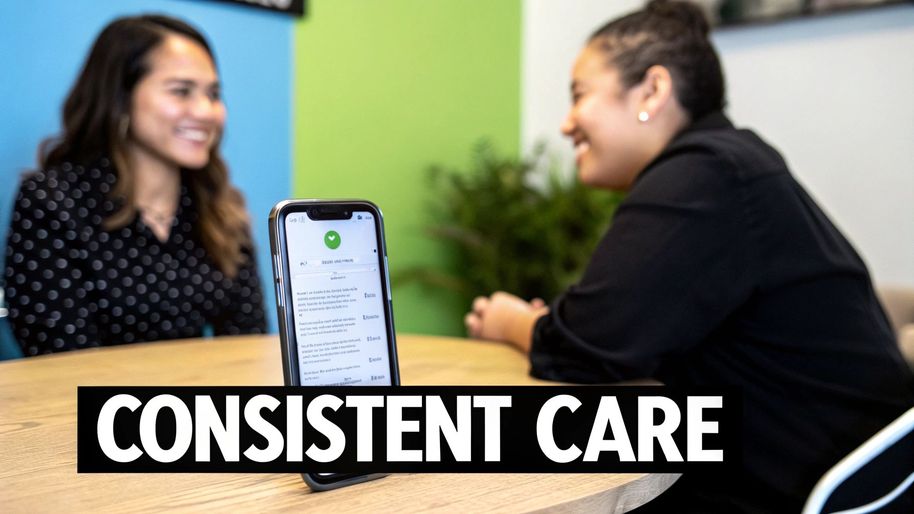 Two women smile while conversing at a table with a smartphone, symbolizing consistent care.