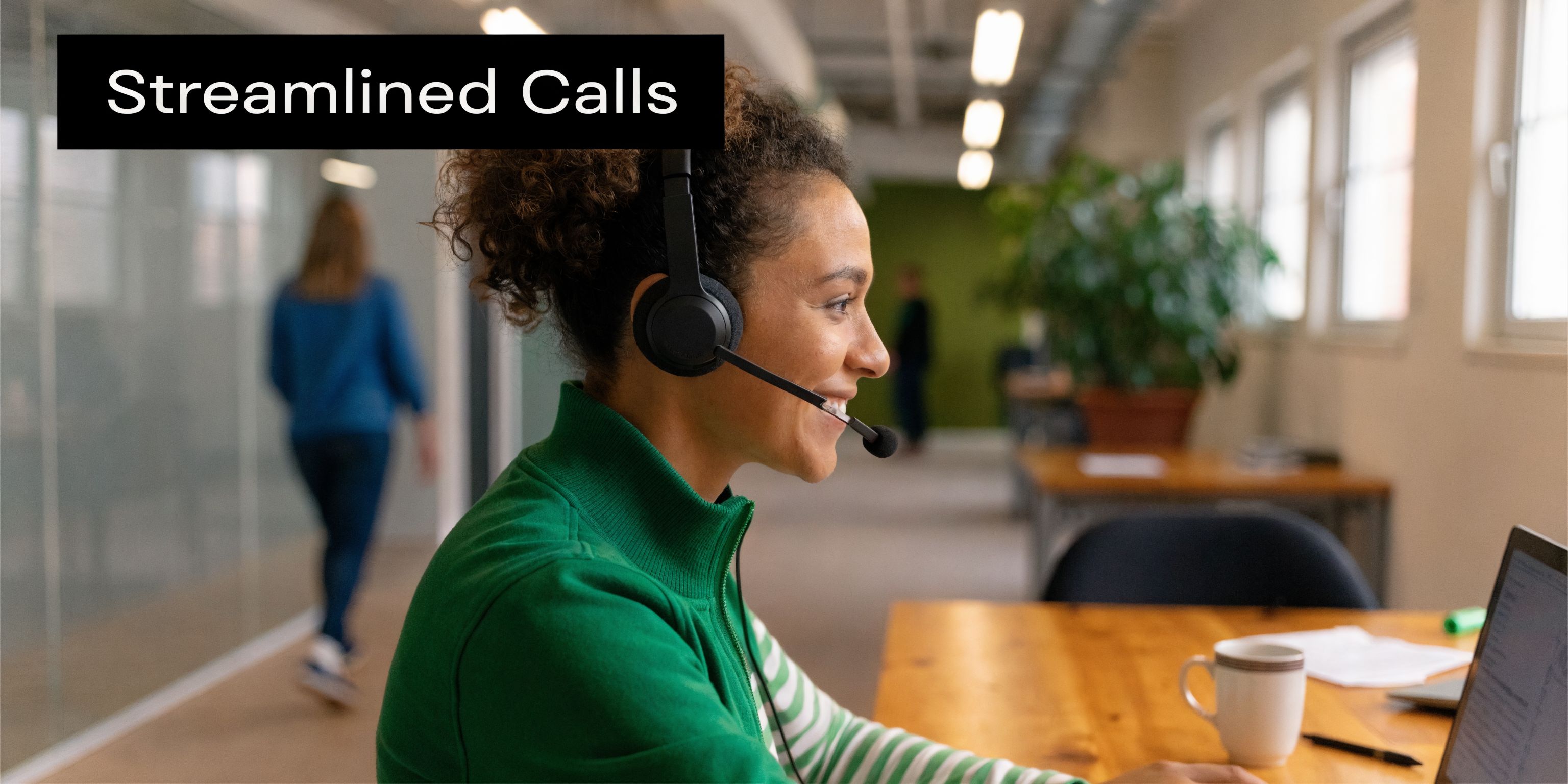 A smiling customer service representative wearing a headset working on a laptop in a bright office.