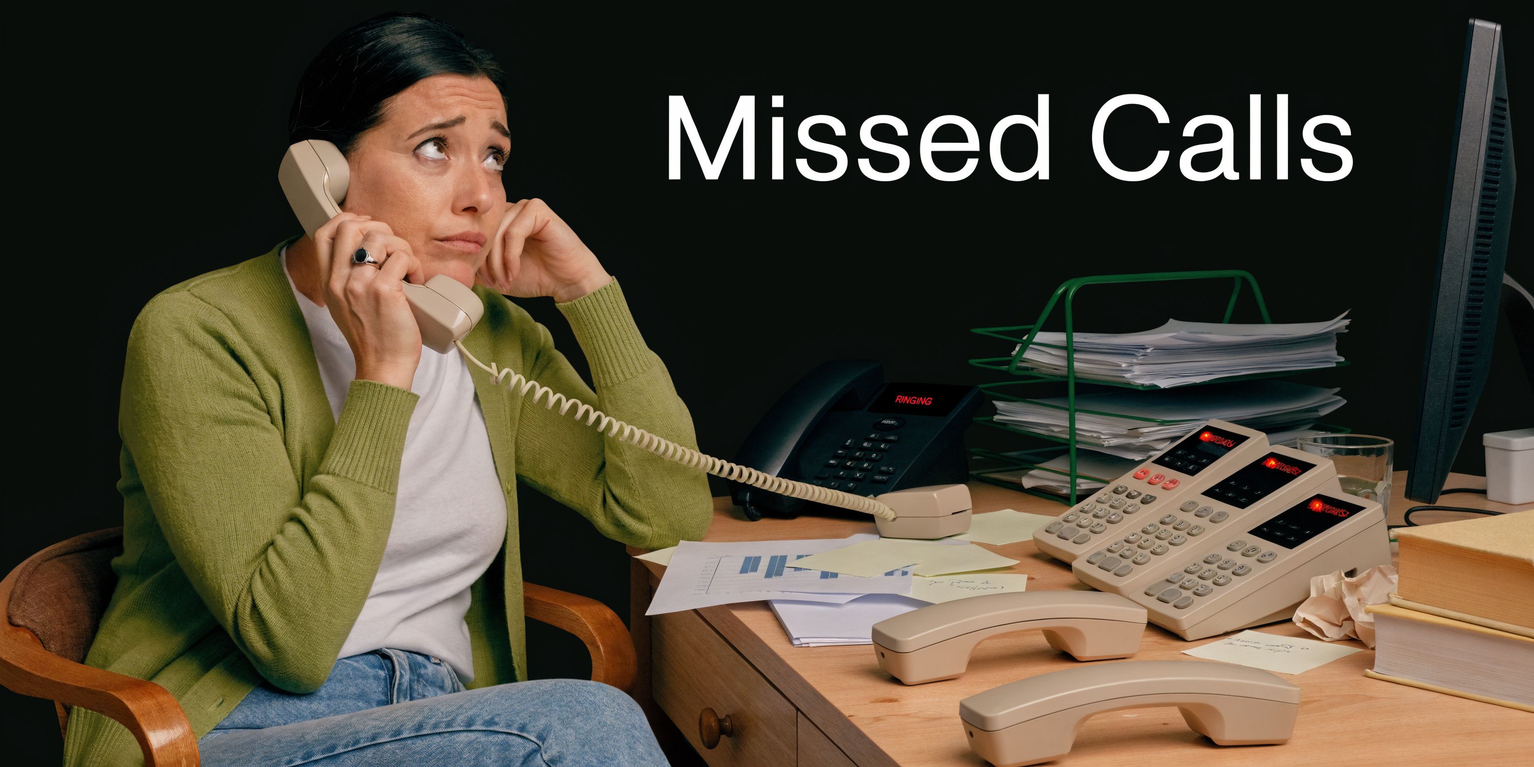 A woman in a green cardigan holds a phone handset while looking stressed in an office setting.
