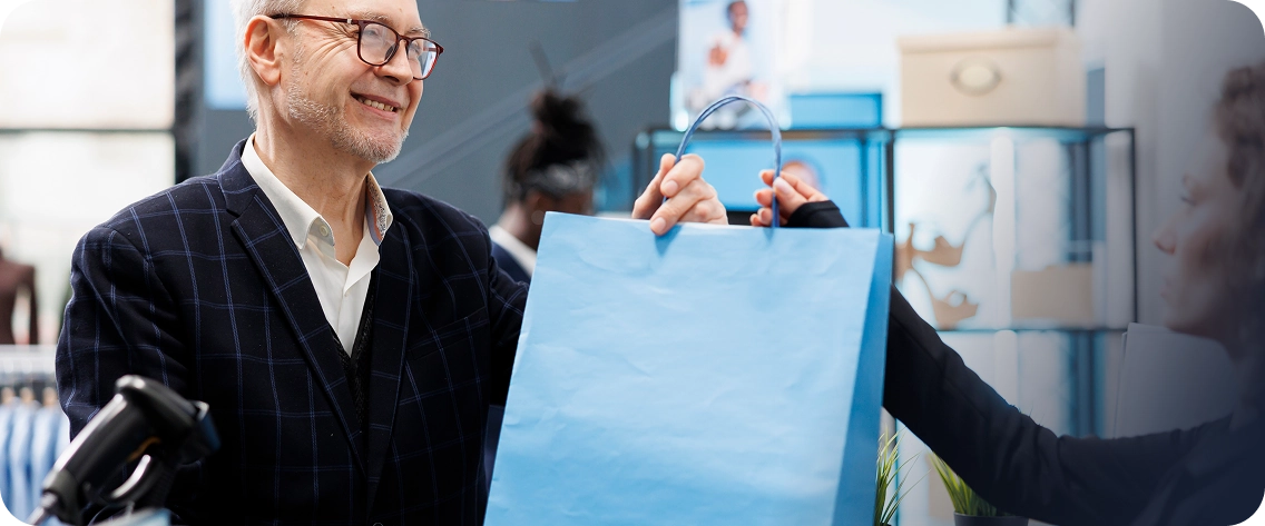 Homem idoso, de cabelo grisalho, óculos e blazer xadrez escuro sobre camisa branca, sorrindo ao receber uma sacola de compras de papel azul claro de uma vendedora em uma loja. A vendedora, parcialmente visível, tem cabelo cacheado e veste uma blusa preta. A cena se passa no caixa, com um leitor de código de barras visível em primeiro plano. O fundo da loja está bem iluminado. Uma sobreposição gráfica azul translúcida está presente no lado direito da imagem.