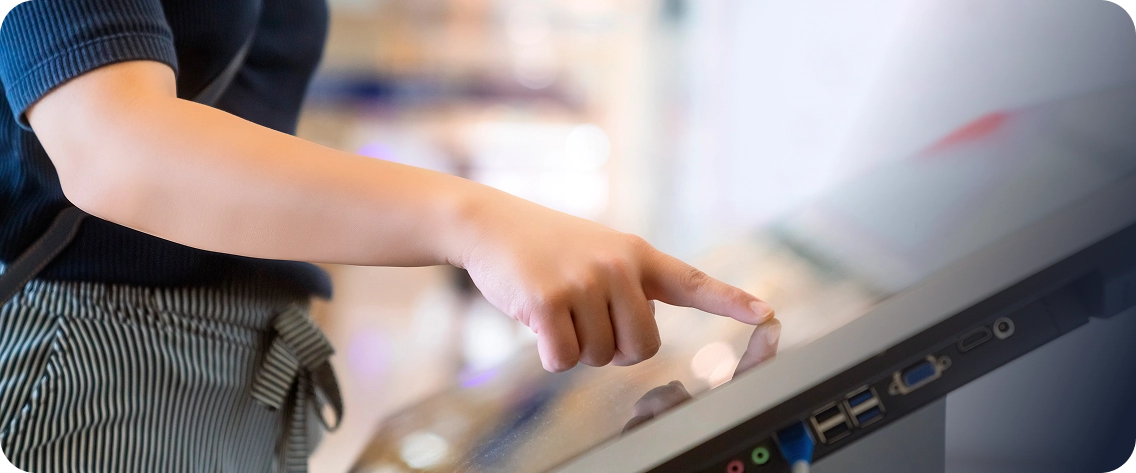 Close-up do braço e da mão de uma pessoa, vestindo blusa escura e calça listrada, que aponta e toca com o dedo indicador em uma grande tela touchscreen inclinada, como a de um terminal de autoatendimento. O fundo está claro e desfocado, sugerindo um ambiente de loja ou espaço público. Uma sobreposição gráfica azul escura e translúcida está presente no lado direito da imagem.