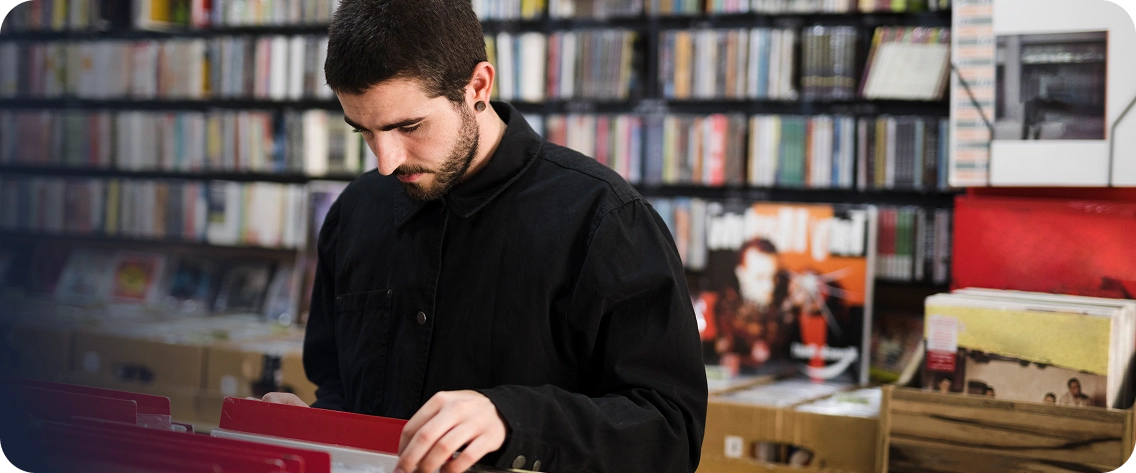 Jovem de barba e camisa preta, com um alargador na orelha, olhando para baixo enquanto vasculha uma caixa de discos de vinil vermelha em uma loja de discos. Ao fundo, estantes altas estão repletas de CDs e vinis. Uma sobreposição gráfica azul escura em gradiente está presente no lado esquerdo da imagem.
