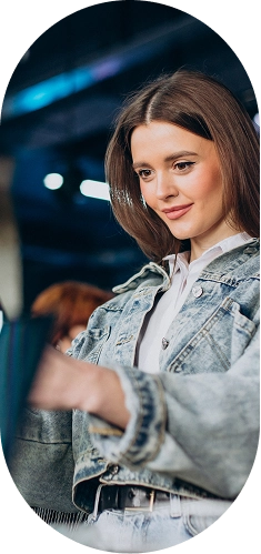 Mulher jovem sorrindo levemente, vestindo uma jaqueta jeans sobre uma camisa branca, olhando para baixo em direção a uma sacola de compras que segura. O fundo está escuro e desfocado, com algumas luzes azuis.