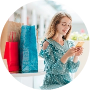 Mulher jovem, loira, sorrindo enquanto usa um smartphone. Ela está sentada ao lado de várias sacolas de compras em uma mesa e veste um vestido azul claro de poá com babados e ombros à mostra. O fundo está claro e desfocado.