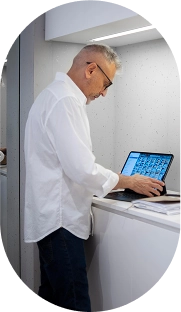 Homem de perfil, com cabelo grisalho curto, óculos e camisa social branca, em pé em um balcão branco. Ele está concentrado, olhando para a tela de um tablet com teclado acoplado e interagindo com ele. O ambiente é claro e moderno. A imagem está contida em uma forma circular com bordas pixelizadas.