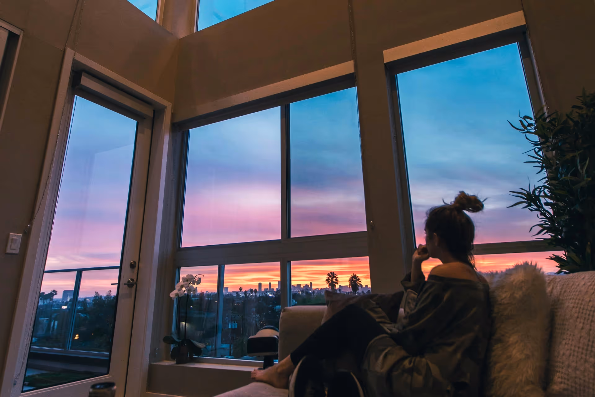 a woman sitting on a couch looking out the windows at a sunset