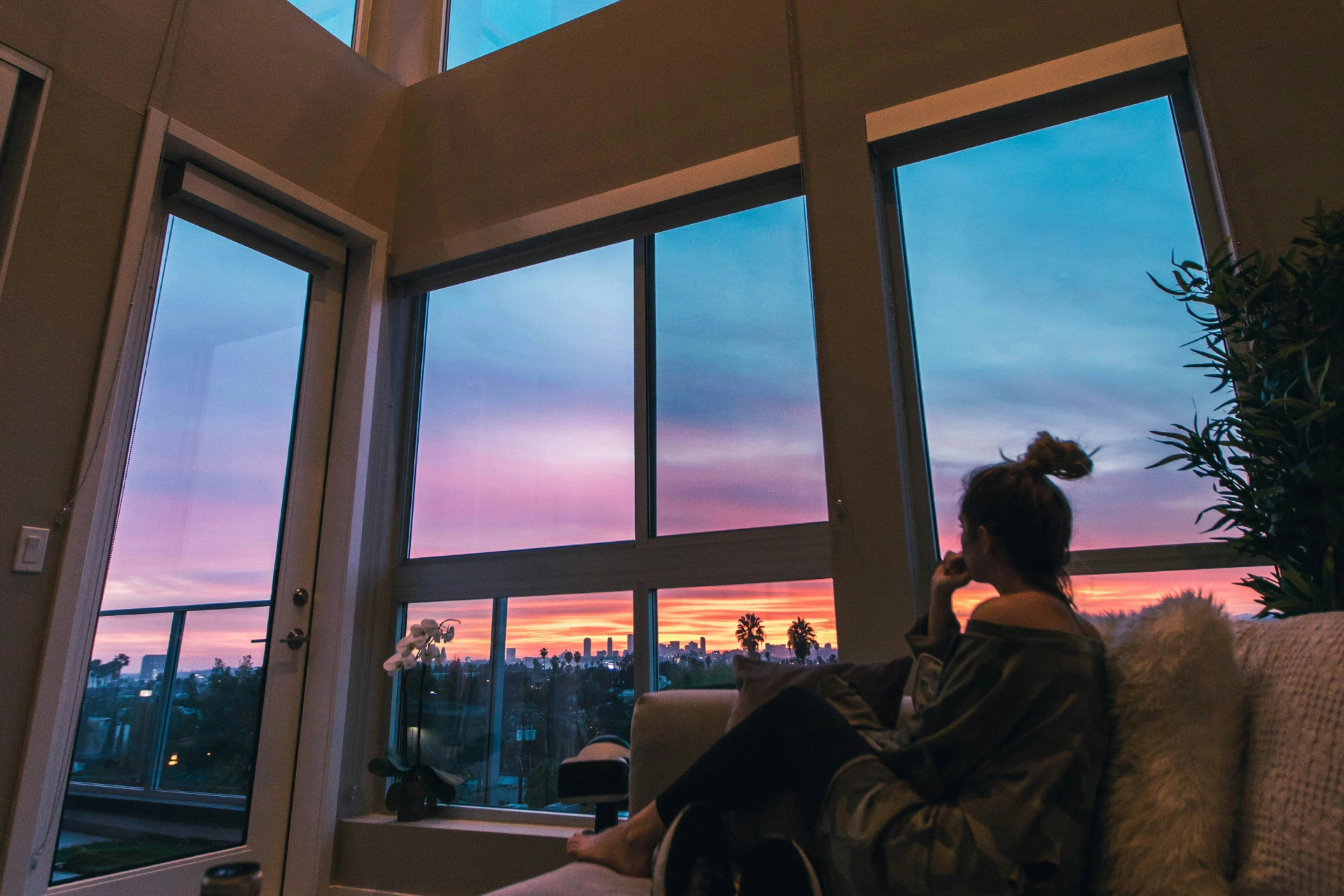 a woman sitting on a couch looking out the windows at a sunset