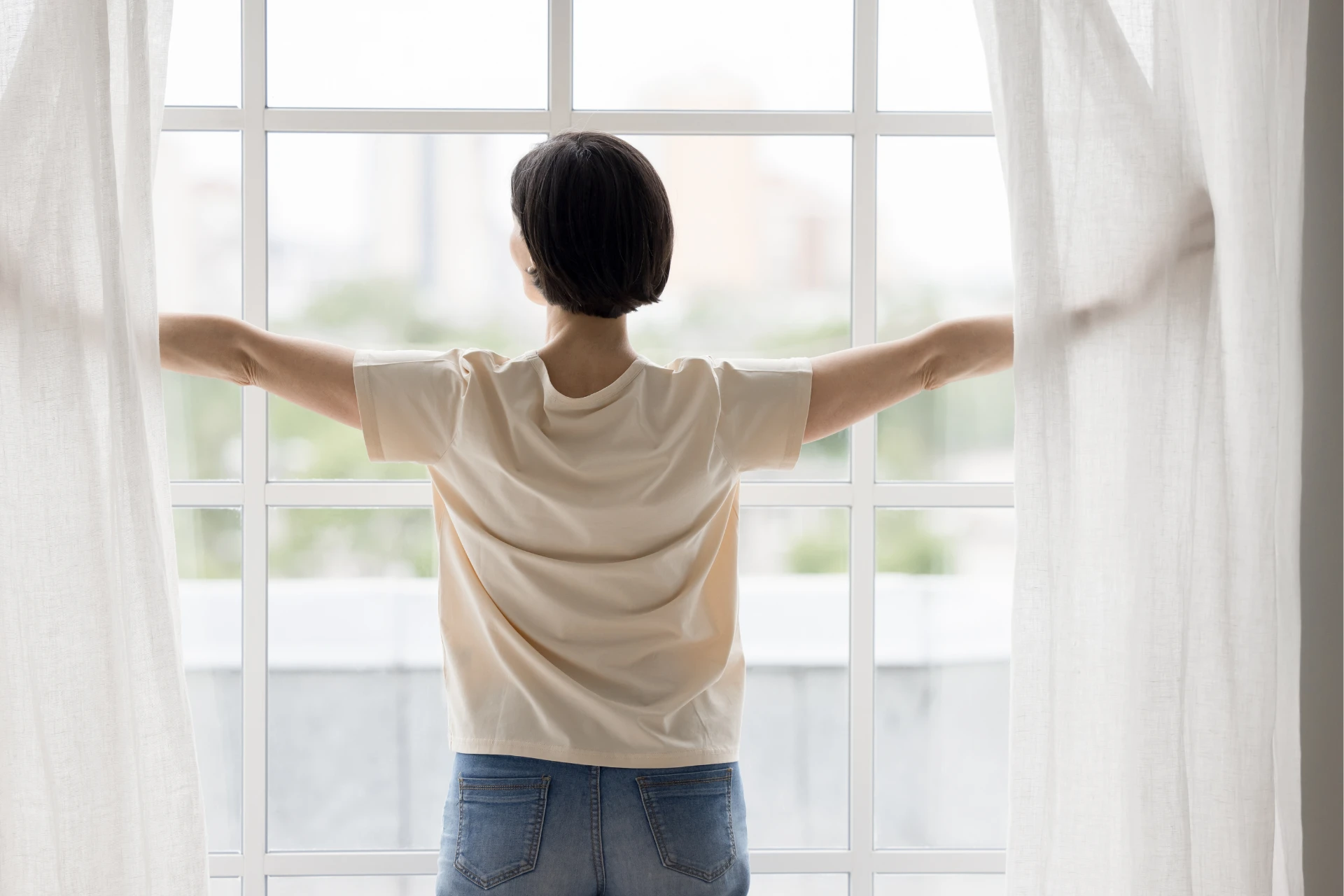 A woman with short, dark hair stands facing away from the viewer, looking out a large, gridded window