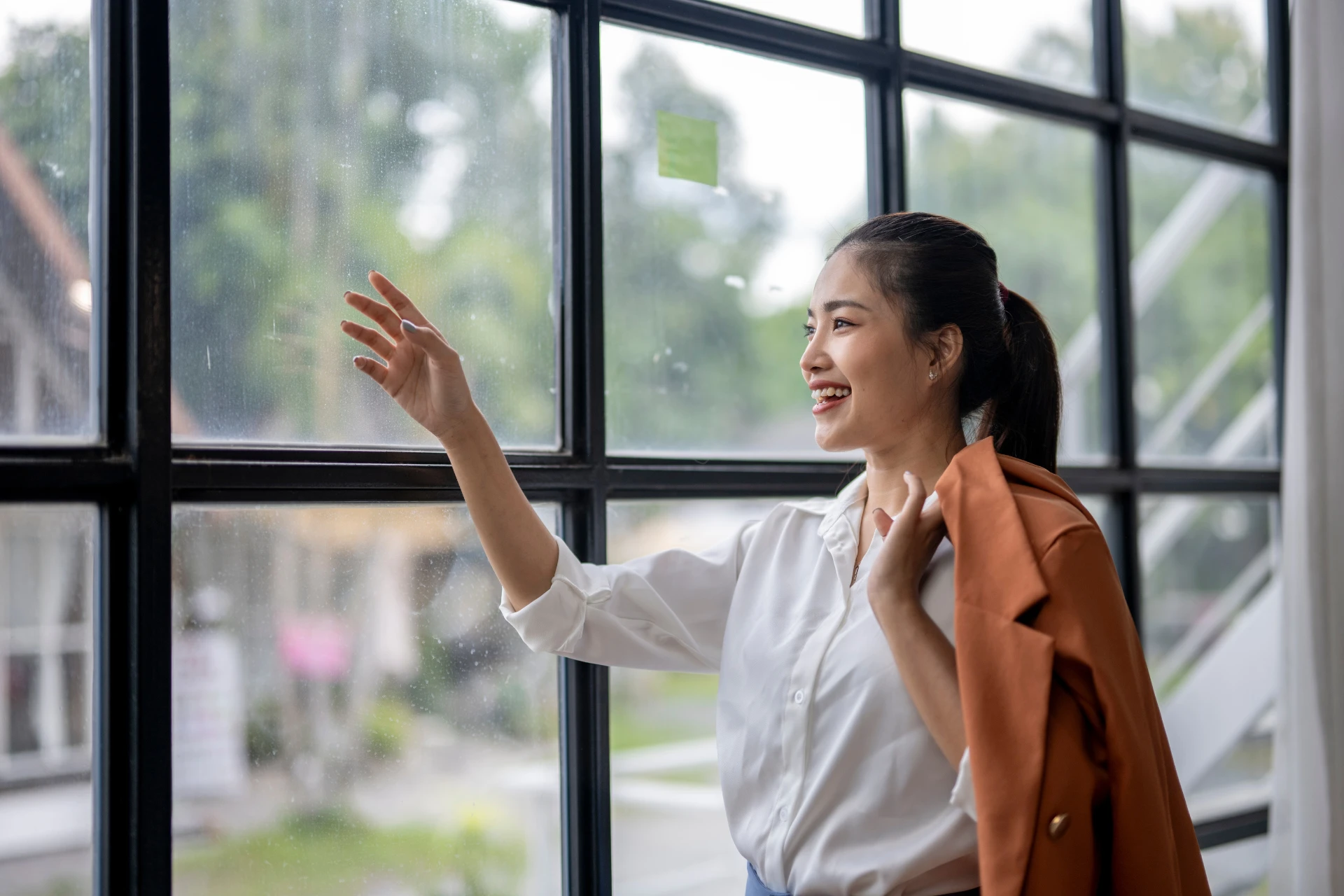 A young woman with her hair pulled back smiles as she leans against a large window