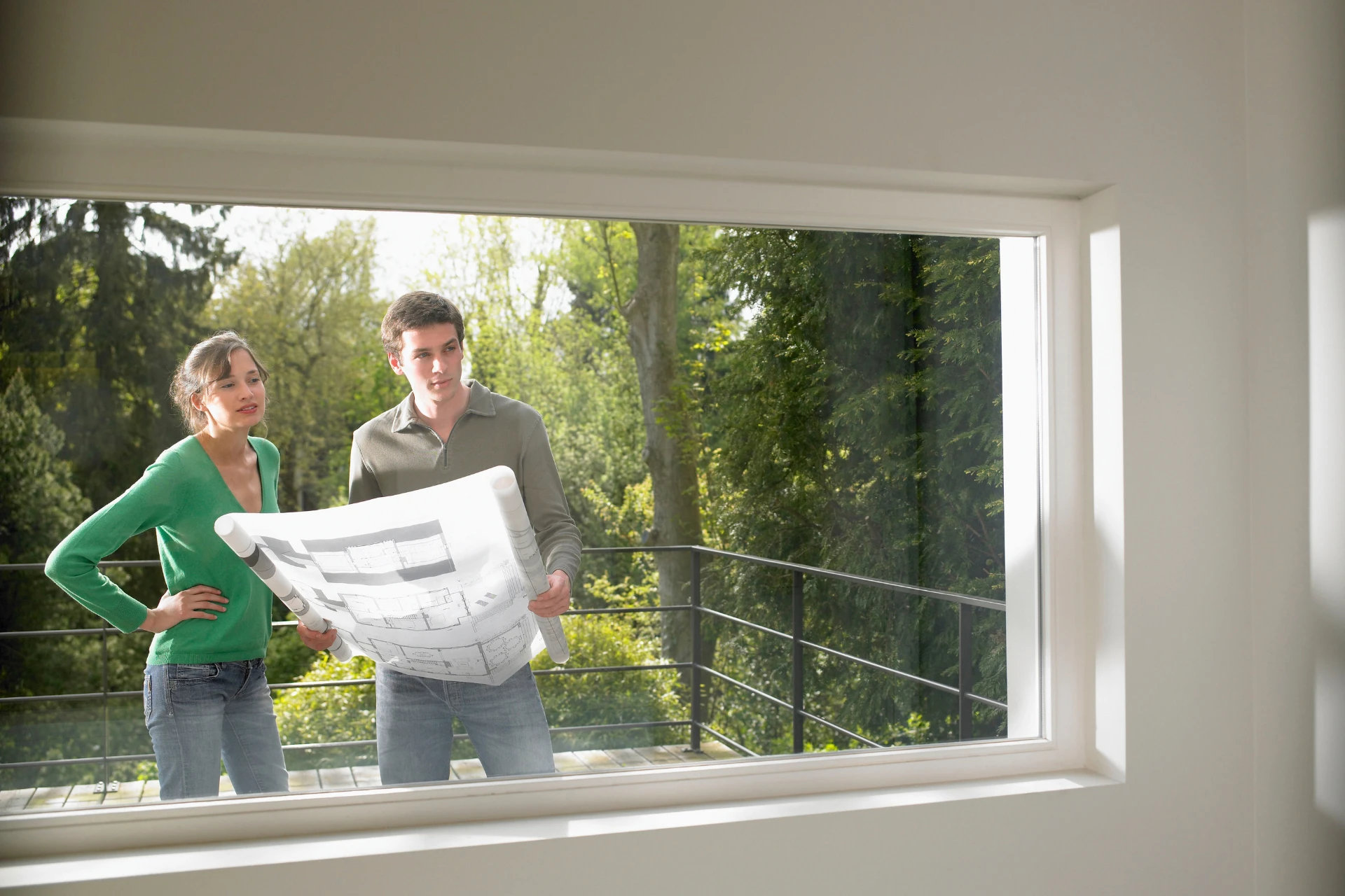 An adult woman and an adult man stand on a balcony, reviewing a large, rolled-out blueprint