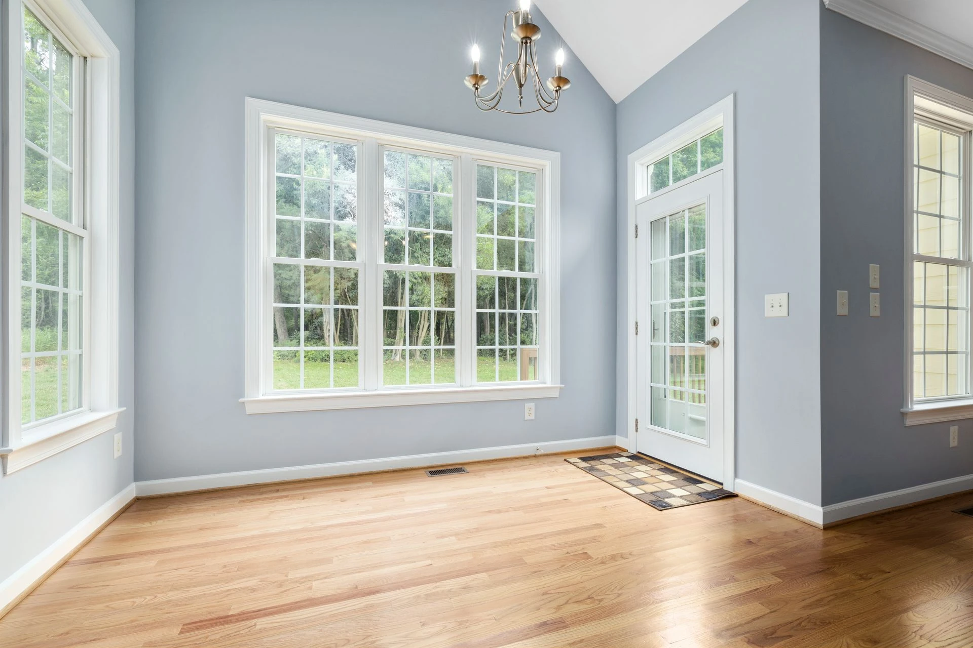Sunlit empty room with wooden floor, chandelier, and French windows leading outside
