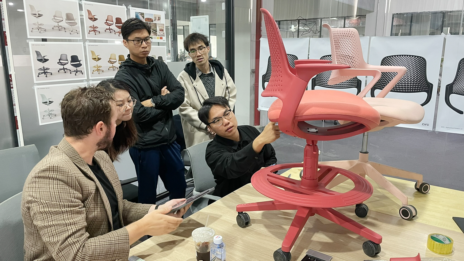 Five people in a meeting room discussing and examining a red office chair prototype on a table.