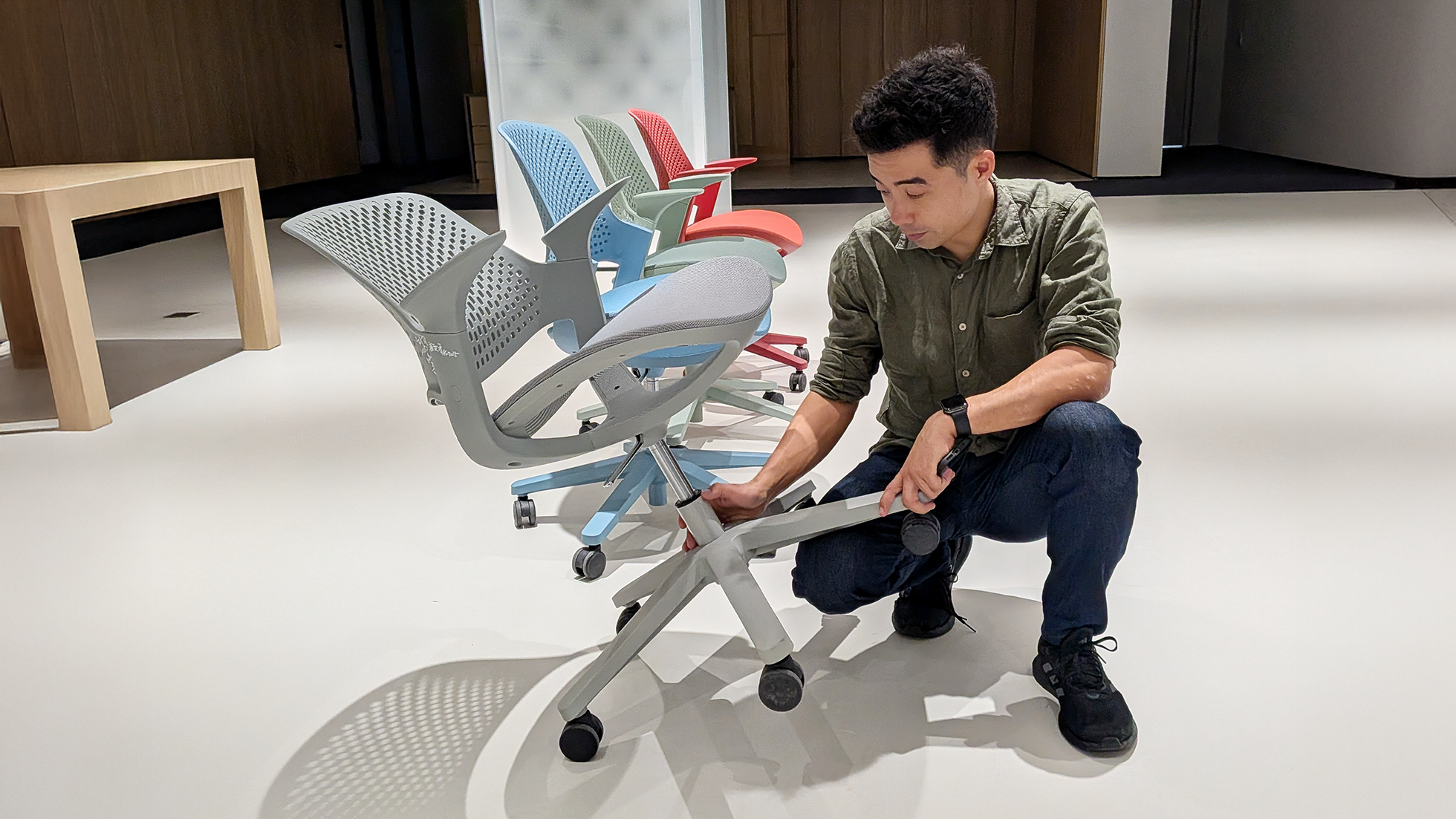 Man in green shirt and jeans kneeling on floor adjusting the wheel of a gray office chair, with three colorful office chairs and a wooden table in the background.
