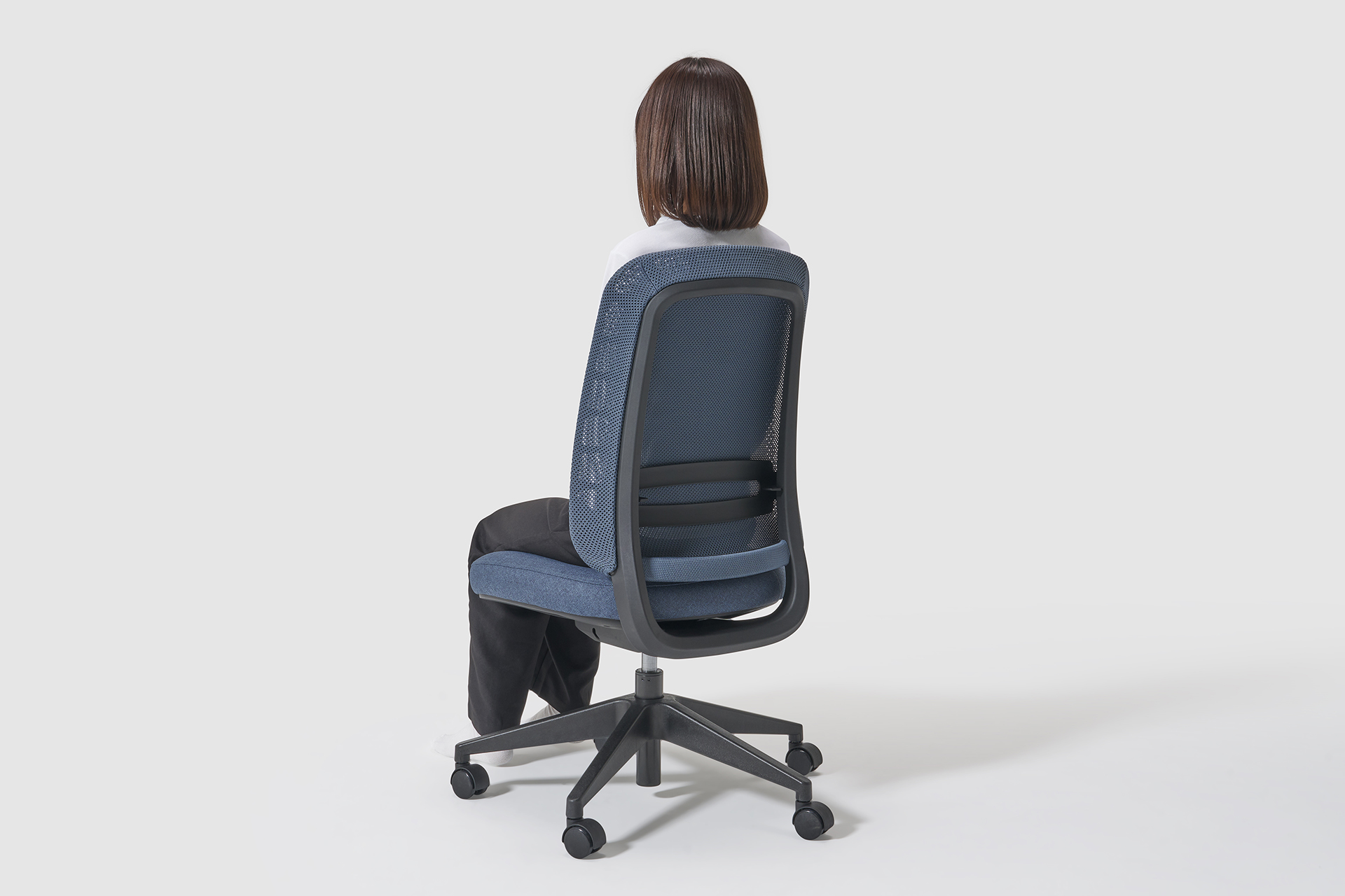A woman sits on a blue mesh back ergonomic chair, facing away from the camera