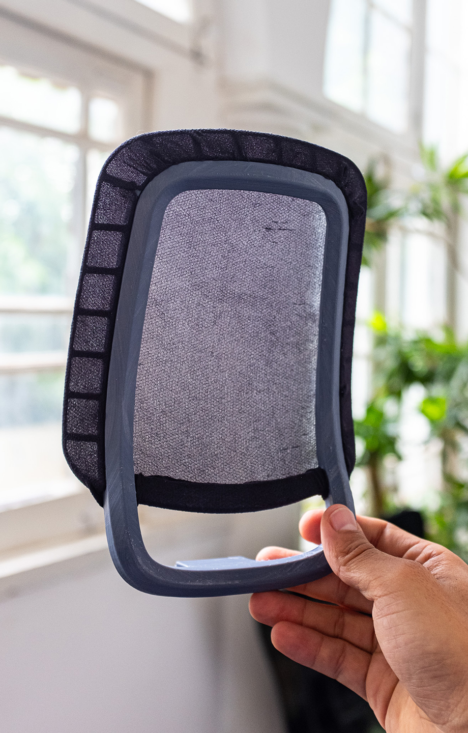 A 1/4 scale prototype of the Mari chair backrest is held up to the window to show the transparency of the mesh and the internal structure