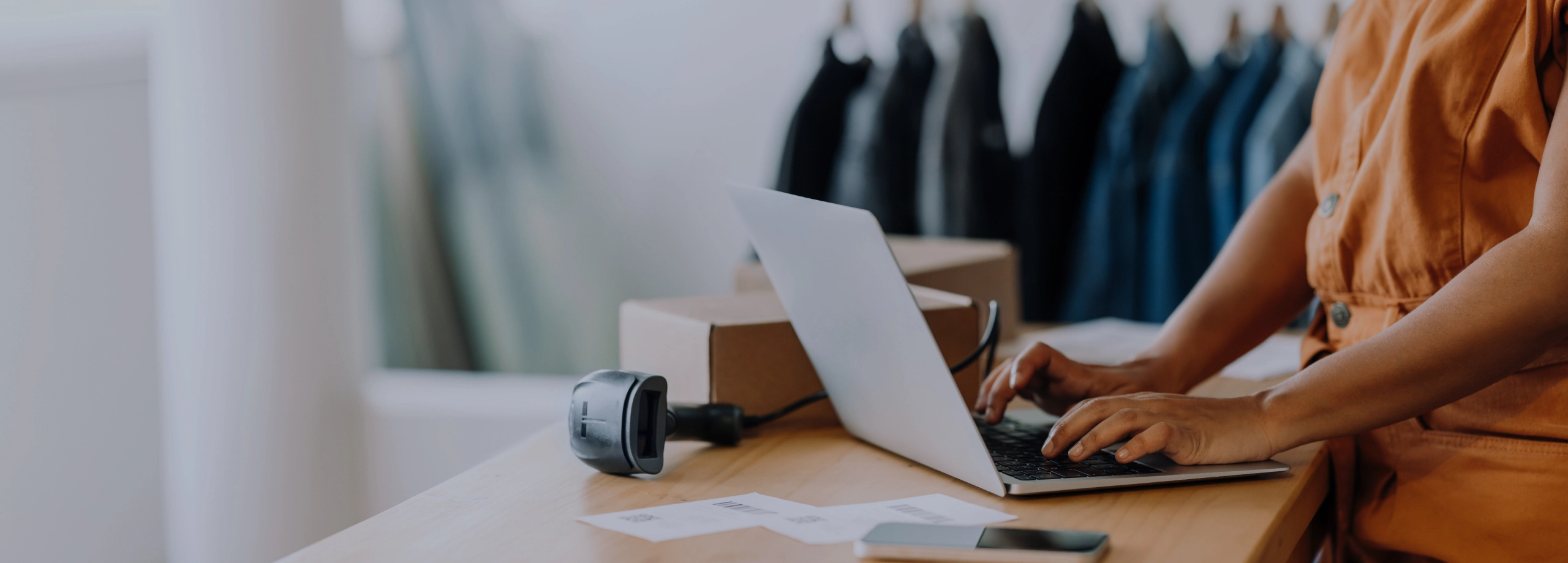 Uma pessoa, possivelmente a dona de um pequeno negócio, está em pé em um balcão de madeira e utiliza um laptop. Na mesa, há também um leitor de código de barras, um smartphone, uma caixa de papelão e papéis, indicando o gerenciamento de um pedido ou inventário. Ao fundo, em um ambiente de loja, há uma arara com roupas penduradas de forma desfocada.