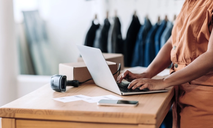 Uma pessoa, possivelmente a dona de um pequeno negócio, está em pé em um balcão de madeira e utiliza um laptop. Na mesa, há também um leitor de código de barras, um smartphone, uma caixa de papelão e papéis, indicando o gerenciamento de um pedido ou inventário. Ao fundo, em um ambiente de loja, há uma arara com roupas penduradas de forma desfocada.