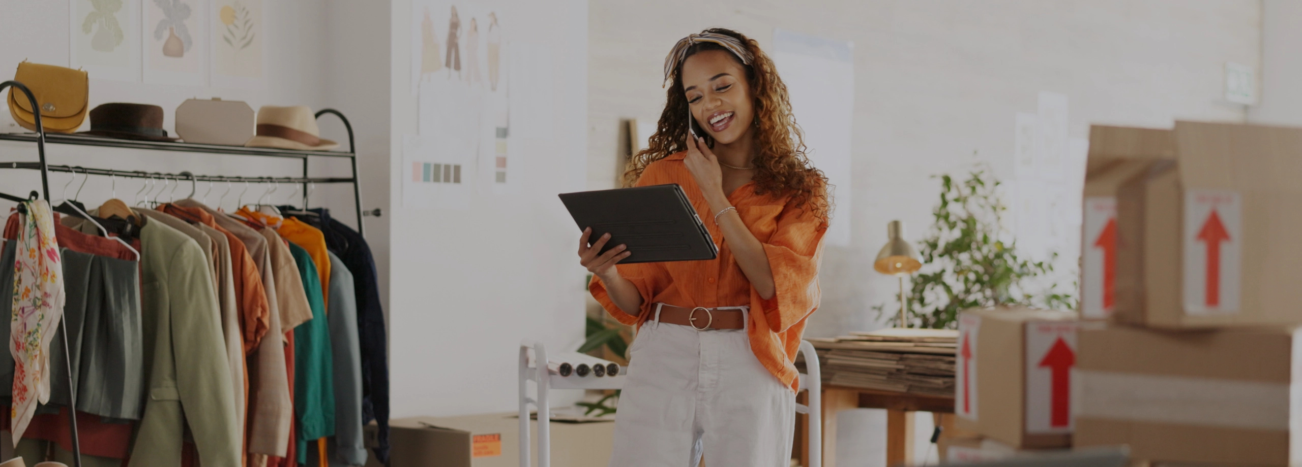 Uma jovem empreendedora de moda, sorrindo, enquanto fala ao celular e olha para um tablet que segura na outra mão. Ela está em pé em seu escritório ou ateliê, vestindo uma camisa laranja e calça branca. Ao fundo, há uma arara com roupas coloridas, chapéus e caixas de papelão prontas para envio, sugerindo um ambiente de e-commerce.