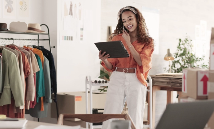 Uma jovem empreendedora de moda, sorrindo, enquanto fala ao celular e olha para um tablet que segura na outra mão. Ela está em pé em seu escritório ou ateliê, vestindo uma camisa laranja e calça branca. Ao fundo, há uma arara com roupas coloridas, chapéus e caixas de papelão prontas para envio, sugerindo um ambiente de e-commerce.