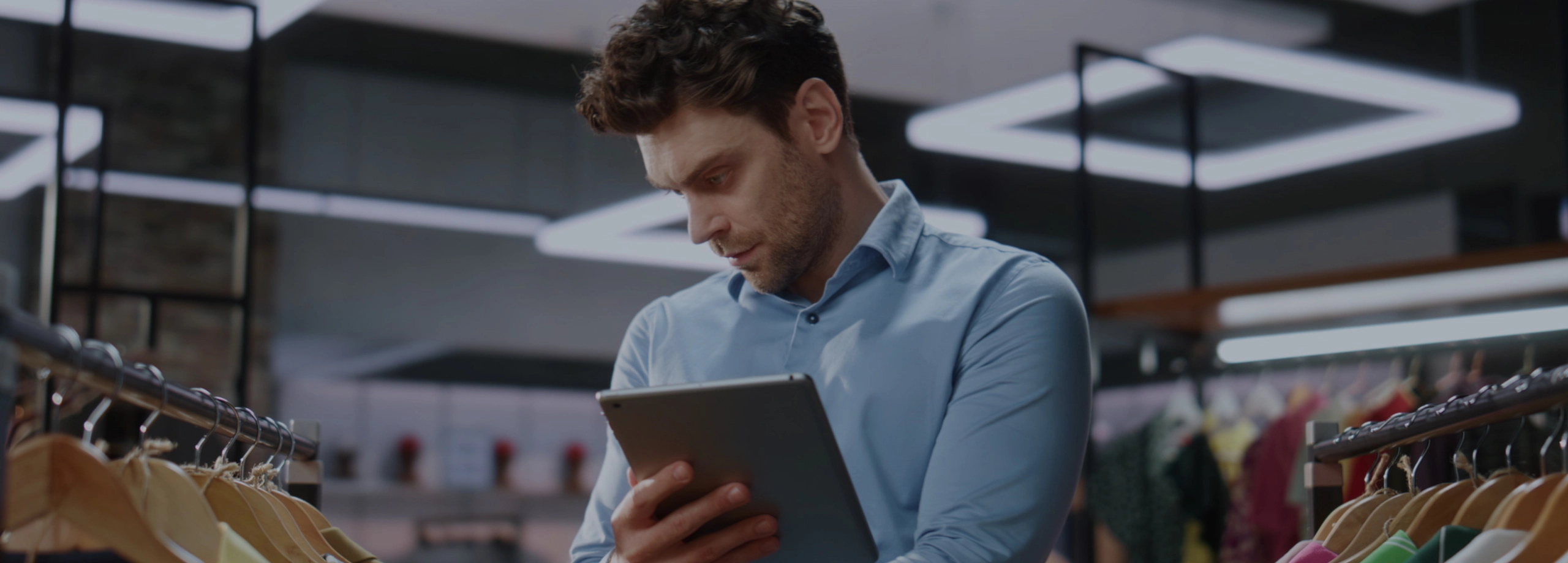 Um homem de camisa azul, possivelmente gerente ou funcionário de uma loja, está em pé em meio a araras de roupas. Ele olha com concentração para um tablet que segura em suas mãos, enquanto verifica o estoque de peças coloridas penduradas em cabides.