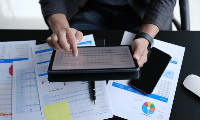 Um homem utilizando um tablet, está sentado em frente a uma mesa e nessa há várias folhas papeis com gráficos imprimidos nelas