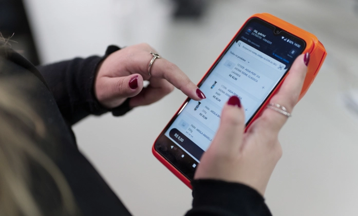 Close-up das mãos de uma pessoa segurando e interagindo com um smartphone com capa laranja. A pessoa está usando o dedo indicador para navegar por um aplicativo de vendas ou estoque que exibe uma lista de produtos, seus códigos e preços. As unhas da pessoa estão pintadas de vermelho e ela usa anéis nos dedos. O foco está no smartphone e na interação, com o fundo levemente desfocado.