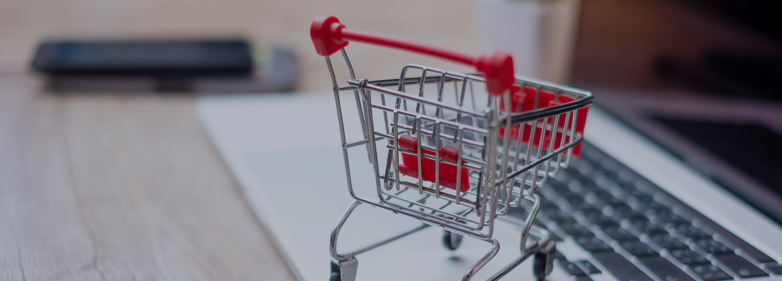Um close-up de um carrinho de compras em miniatura, feito de metal prateado e plástico rosa, que está parado em cima de um laptop aberto. O carrinho está ligeiramente inclinado para a direita e seus detalhes, como as rodinhas e a alça, estão nítidos. O teclado do laptop está visível abaixo do carrinho, e a tela está fora de foco ao fundo, com um leve brilho. O plano de fundo é desfocado, sugerindo um ambiente de escritório ou doméstico.