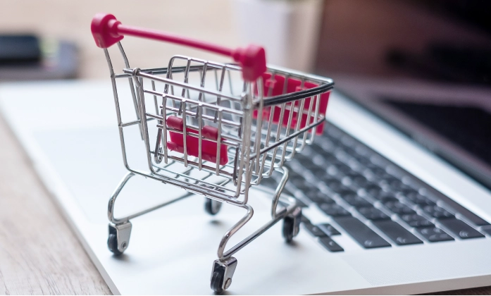 Um close-up de um carrinho de compras em miniatura, feito de metal prateado e plástico rosa, que está parado em cima de um laptop aberto. O carrinho está ligeiramente inclinado para a direita e seus detalhes, como as rodinhas e a alça, estão nítidos. O teclado do laptop está visível abaixo do carrinho, e a tela está fora de foco ao fundo, com um leve brilho. O plano de fundo é desfocado, sugerindo um ambiente de escritório ou doméstico.