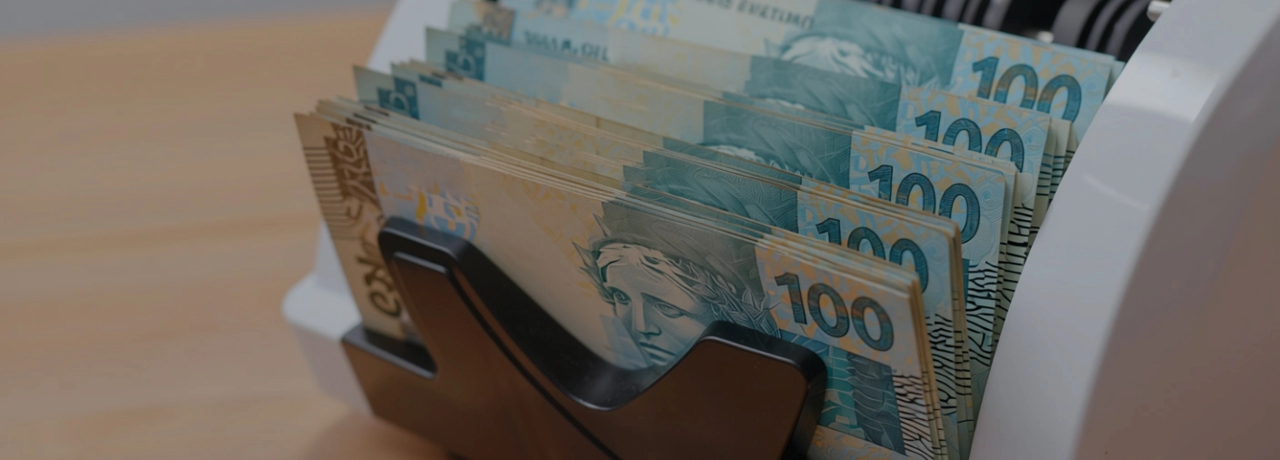 Um close-up de várias notas de 100 Reais sendo processadas por uma máquina de contar dinheiro. As notas, predominantemente azuis e beges, estão alinhadas e empilhadas na abertura da máquina, com a Efígie da República visível em algumas delas. A máquina, de cor clara (branca ou creme), ocupa o lado direito da imagem e parte superior, e os rolos internos pretos da máquina são visíveis ao fundo, sugerindo o movimento de contagem. O fundo à esquerda é uma superfície de madeira clara e desfocada.