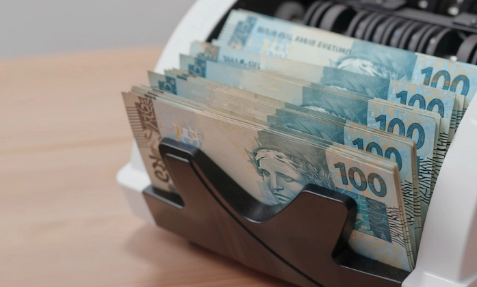 Um close-up de várias notas de 100 Reais sendo processadas por uma máquina de contar dinheiro. As notas, predominantemente azuis e beges, estão alinhadas e empilhadas na abertura da máquina, com a Efígie da República visível em algumas delas. A máquina, de cor clara (branca ou creme), ocupa o lado direito da imagem e parte superior, e os rolos internos pretos da máquina são visíveis ao fundo, sugerindo o movimento de contagem. O fundo à esquerda é uma superfície de madeira clara e desfocada.