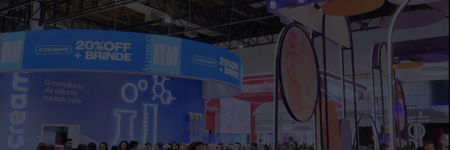 Uma jovem mulher com cabelos castanhos longos e lisos está de pé no centro de um palco, sorrindo e olhando diretamente para a câmera. Ela veste uma blusa preta sem mangas e uma saia preta com um cinto fino. Com uma das mãos, ela segura um microfone perto da boca, e a outra mão está levantada, com a palma aberta, como se estivesse acenando ou gesticulando. Atrás dela, há um fundo iluminado e translúcido em tons de azul e laranja, com um design abstrato que inclui formas circulares e retangulares, reminiscentes de um estande de feira. Garrafas de produtos de beleza com etiquetas coloridas são visíveis através do fundo translúcido, sugerindo um evento relacionado à beleza. Um retângulo transparente sobrepõe a imagem, com a frase "Creamy na Beauty fair 2025 com a Nexaas" escrita em branco, dividida em dois blocos de texto. No canto superior central, há um logo branco com um "N" estilizado e a palavra "Nexaas" em preto, sobre um fundo branco retangular.