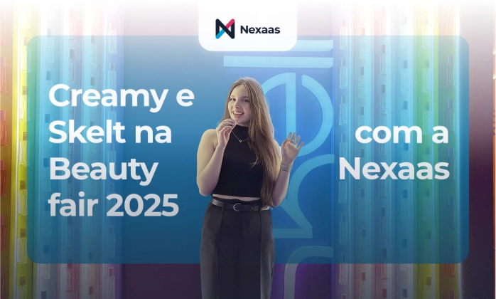 Uma jovem mulher com cabelos castanhos longos e lisos está de pé no centro de um palco, sorrindo e olhando diretamente para a câmera. Ela veste uma blusa preta sem mangas e uma saia preta com um cinto fino. Com uma das mãos, ela segura um microfone perto da boca, e a outra mão está levantada, com a palma aberta, como se estivesse acenando ou gesticulando. Atrás dela, há um fundo iluminado e translúcido em tons de azul e laranja, com um design abstrato que inclui formas circulares e retangulares, reminiscentes de um estande de feira. Garrafas de produtos de beleza com etiquetas coloridas são visíveis através do fundo translúcido, sugerindo um evento relacionado à beleza. Um retângulo transparente sobrepõe a imagem, com a frase "Creamy na Beauty fair 2025 com a Nexaas" escrita em branco, dividida em dois blocos de texto. No canto superior central, há um logo branco com um "N" estilizado e a palavra "Nexaas" em preto, sobre um fundo branco retangular.
