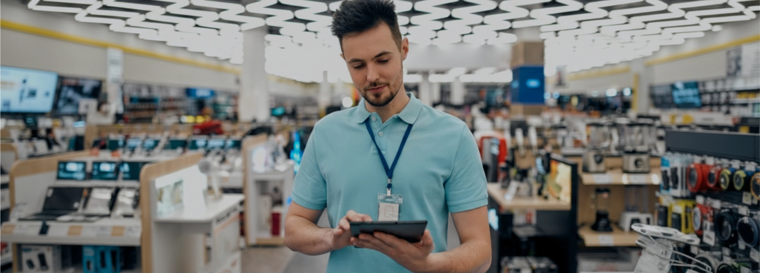 Um jovem vendedor em uma loja de eletrônicos, vestindo uma camisa polo azul clara e um crachá de identificação, está olhando para baixo e interagindo com um tablet digital que ele segura com as duas mãos. Ele tem uma barba curta e bem cuidada. O fundo da imagem está desfocado, mostrando prateleiras com produtos eletrônicos e iluminação moderna no teto da loja.