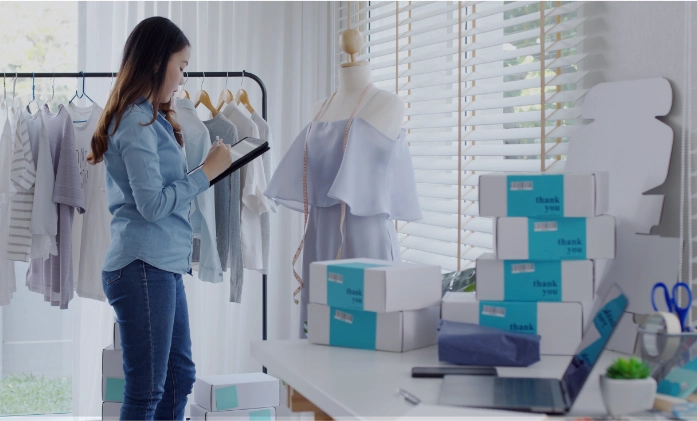 Mulher em pé segura um tablet e confere roupas em uma arara em um ateliê de moda, com um manequim e caixas de encomenda sobre uma mesa ao lado.