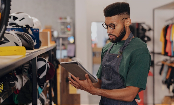 Funcionário de loja de bicicletas, vestindo avental e óculos, consulta um tablet em frente a uma prateleira com capacetes.