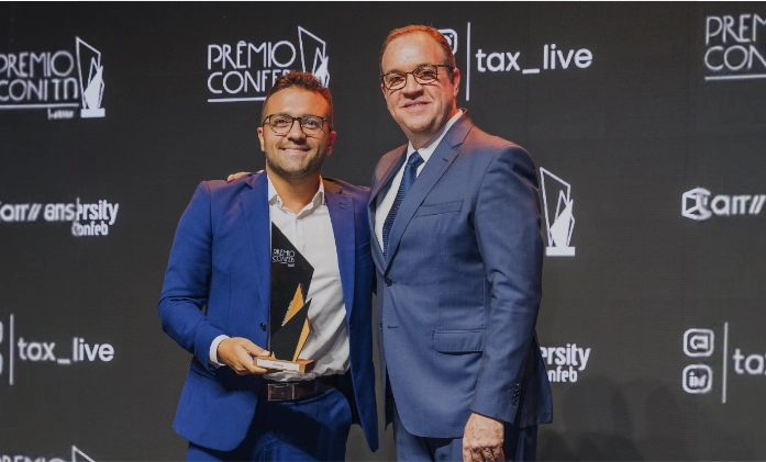Foto de dois homens abraçados em um palco de premiação. O homem à esquerda, de terno azul royal e sem gravata, segura um troféu geométrico preto e dourado. O homem à direita, de terno azul marinho e gravata, sorri ao seu lado. Ao fundo, um painel preto com as logomarcas 'Prêmio Confeb', 'tax_live' e 'Live University'.