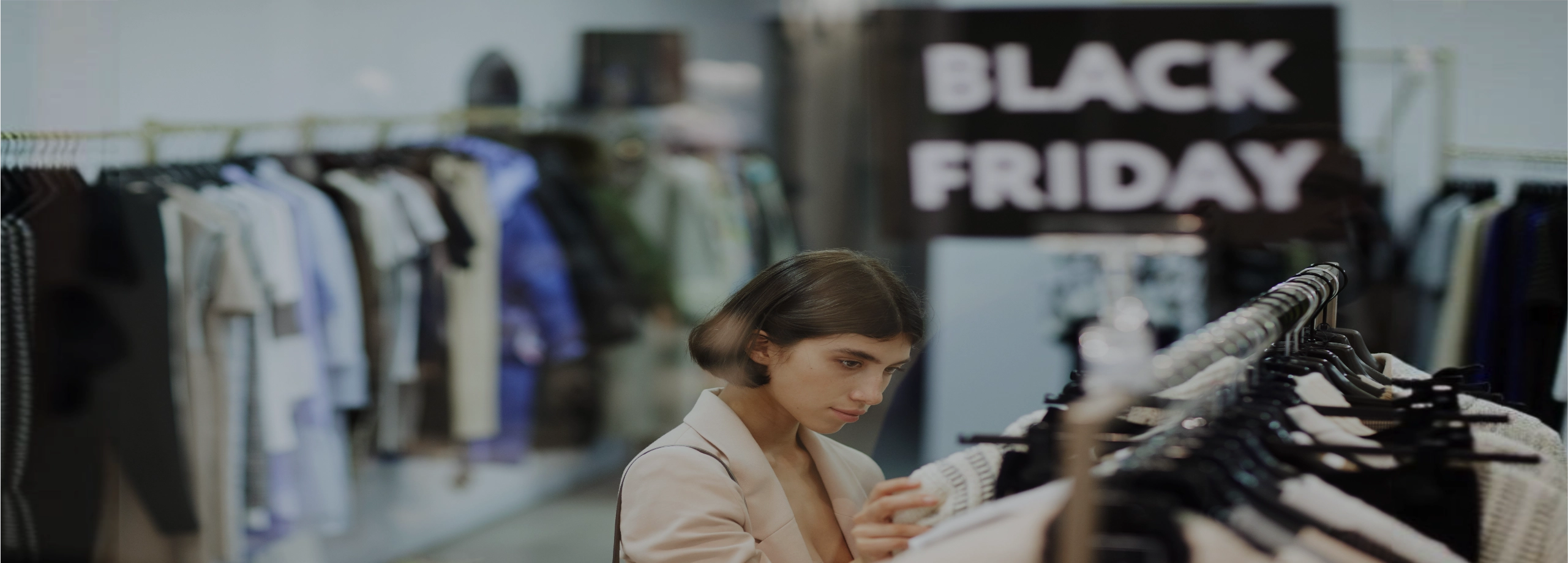 Uma mulher jovem de cabelo curto e blazer bege está focada, segurando e analisando uma roupa branca em um cabide. Ela está em uma loja de moda. Ao fundo, levemente desfocado, vê-se outras araras com roupas e uma placa preta com letras brancas grandes escrito 'BLACK FRIDAY'.