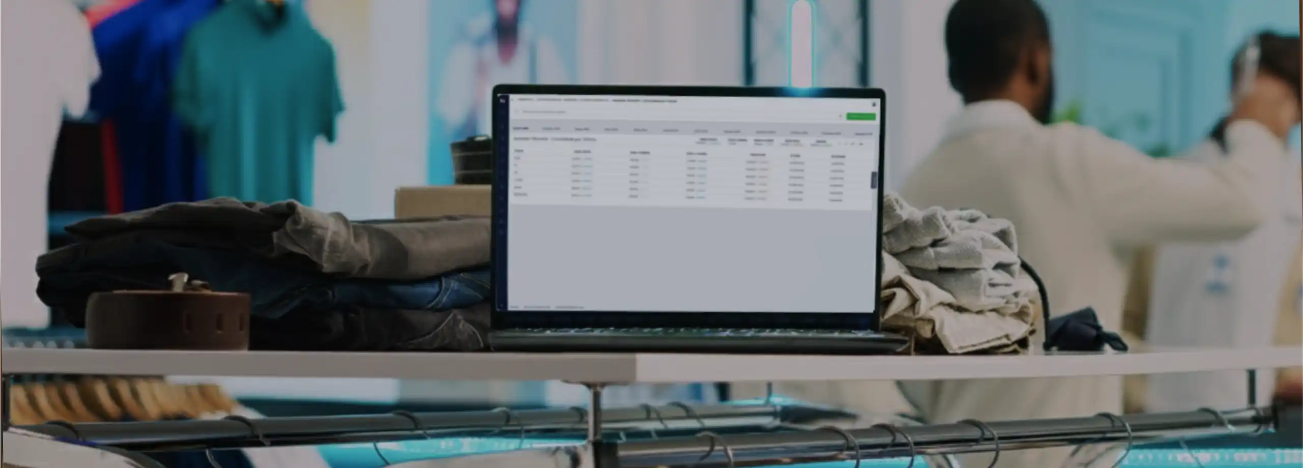 Em primeiro plano, um notebook está apoiado sobre uma prateleira branca, cercado por pilhas de calças jeans e roupas bege dobradas. A tela exibe uma tabela de dados de um software corporativo. Abaixo da prateleira, vê-se o topo de araras metálicas. O fundo está desfocado, mostrando o ambiente da loja com pôsteres de modelos na parede e um cliente, de costas, olhando as roupas.