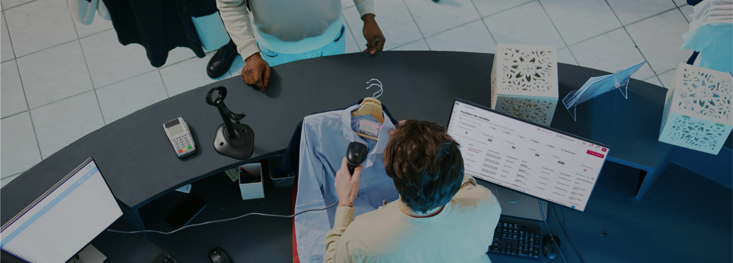 Vista aérea de um caixa de loja escaneando uma camisa azul para um cliente, com o monitor exibindo o sistema de vendas.