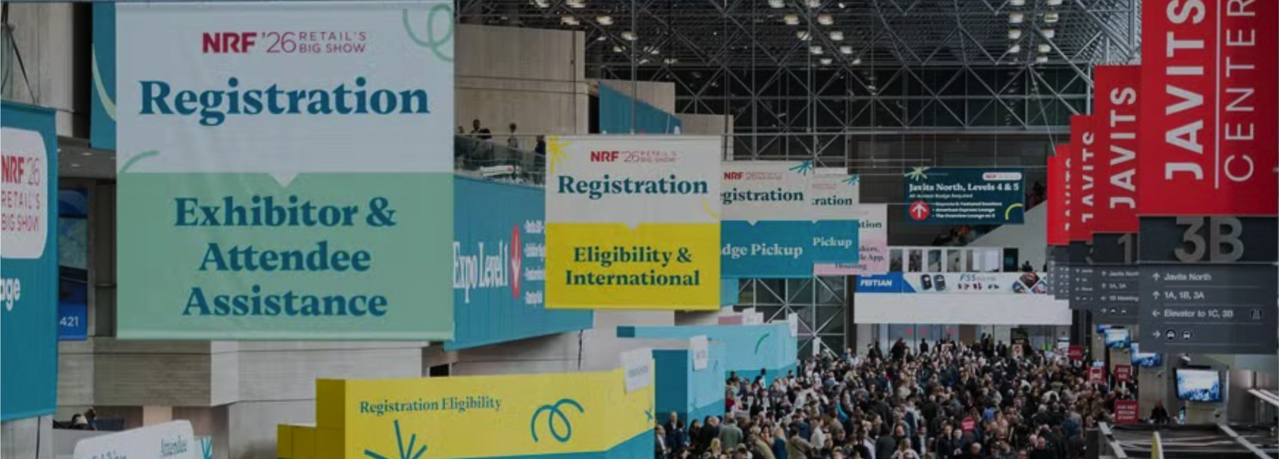 Uma visão ampla do interior do Javits Center repleto de milhares de participantes. No teto alto, pendem vários banners grandes de sinalização para o evento 'NRF '26 Retail's Big Show'. Um banner verde indica 'Registration: Exhibitor & Attendee Assistance' e um amarelo indica 'Eligibility & International'. À direita, faixas verticais vermelhas mostram o nome 'JAVITS CENTER'.